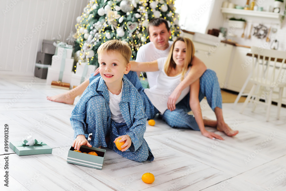 Fototapeta premium a small cute baby boy in blue pajamas takes tangerines out of a gift box and smiles slyly against the background of his parents and the Christmas tree.