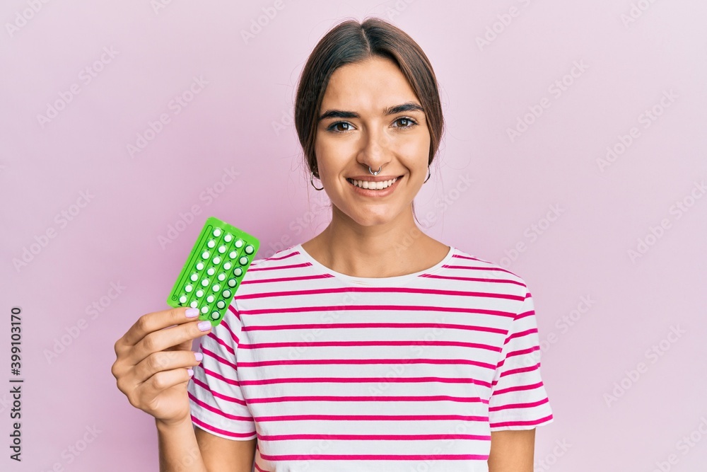 Young hispanic woman holding birth control pills looking positive and happy standing and smiling with a confident smile showing teeth Young hispanic woman holding birth control pills looking positive and happy standing and smiling with a confident smile showing teeth
