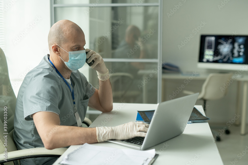 Side view of mature male clinician in mask sitting by desk in front of laptop Side view of mature male clinician in mask sitting by desk in front of laptop