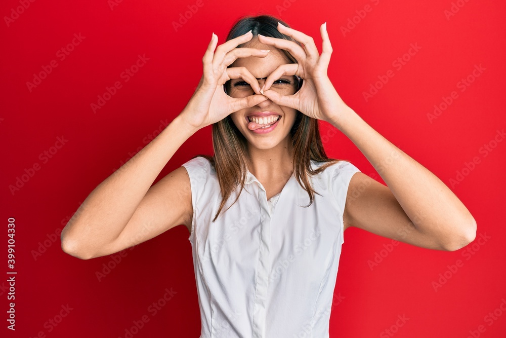 Young brunette woman wearing casual clothes and glasses doing ok gesture like binoculars sticking tongue out, eyes looking through fingers. crazy expression. Young brunette woman wearing casual clothes and glasses doing ok gesture like binoculars sticking tongue out, eyes looking through fingers. crazy expression.
