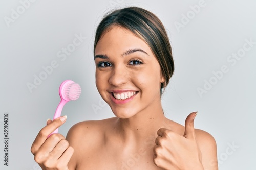 Young brunette woman using facial exfoliating brush smiling happy and positive, thumb up doing excellent and approval sign