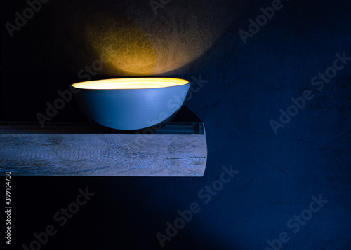 Close-up of a bamboo bowl, on a wooden shelf, glowing yellow light on a dark background.