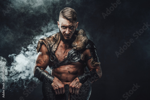 Portrait of evil nordic pagan in light armour with fur staring at camera and holding two handed axe in dark and foggy background.