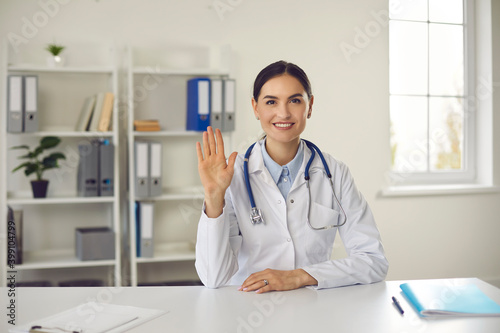 Doctor waving her hand in greeting, giving an online consultation to a patient looking at a webcam
