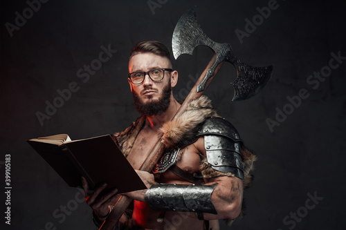 Holding a book scandinavian warrior weared with glasses dressed in light armour with two handed axe in dark background.