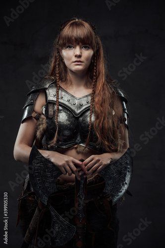 Beautiful but dangerous warlike woman viking with long brown hairs in dark armour holding huge axe in dark background.