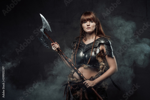 Portrait of warlike female fighter from north with long brown hairs in dark armour with two handed axe in dark smokey background.