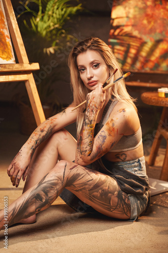 Portrait of creative hipster woman with blond hairs and tattooed leg and arm holding paintbrushes in shiny and warm workshop.
