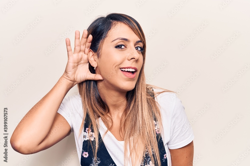Young beautiful woman wearing casual floral dress smiling with hand over ear listening and hearing to rumor or gossip. deafness concept. Young beautiful woman wearing casual floral dress smiling with hand over ear listening and hearing to rumor or gossip. deafness concept.