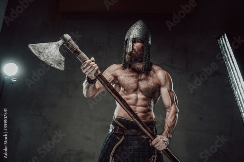 Muscular and furious nord warrior staying with naked torso with helmet on his head holding his two handed axe in dark background.