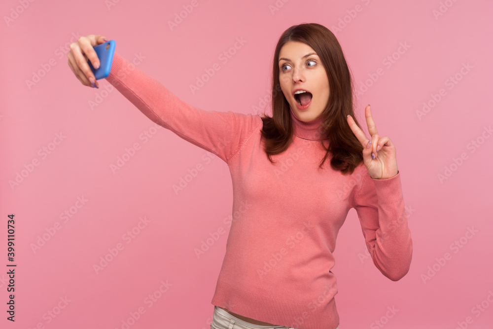 Funny female blogger with brown hair showing peace gesture and fooling face recording video or making photo on smartphone. Indoor studio shot isolated on pink background Funny female blogger with brown hair showing peace gesture and fooling face recording video or making photo on smartphone. Indoor studio shot isolated on pink background