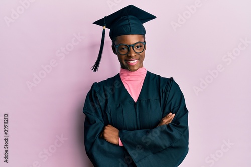 Young african american girl wearing graduation cap and ceremony robe happy face smiling with crossed arms looking at the camera. positive person.
