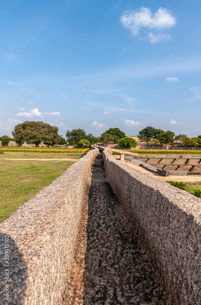 Hampi, Karnataka, India - November 4, 2013: Royal Enclosure. Long view on dry brown stone aqueduct through green garden and passing to stepped tank under blue cloudscape. Hampi, Karnataka, India - November 4, 2013: Royal Enclosure. Long view on dry brown stone aqueduct through green garden and passing to stepped tank under blue cloudscape.