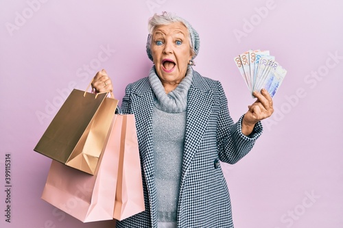 Senior grey-haired woman holding shopping bags and swedish krona banknotes celebrating crazy and amazed for success with open eyes screaming excited.