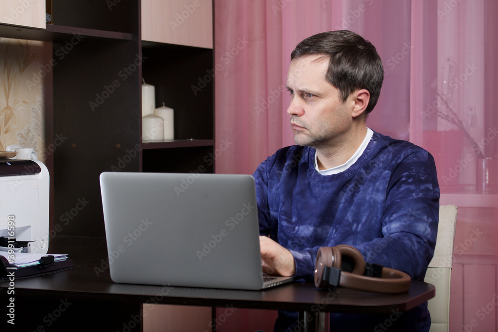 A man in home clothes works at a laptop. Headphones are on the table nearby. A man in home clothes works at a laptop. Headphones are on the table nearby.