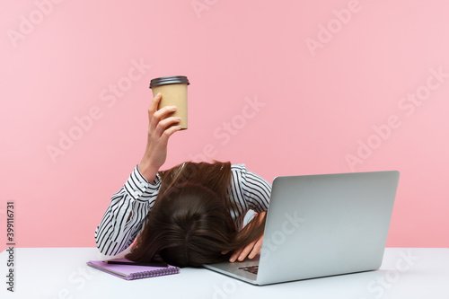 Obraz na plátne Sleepy bored woman office worker lying on table with laptop holding and showing paper coffee cup, feeling lack of energy sitting at workplace