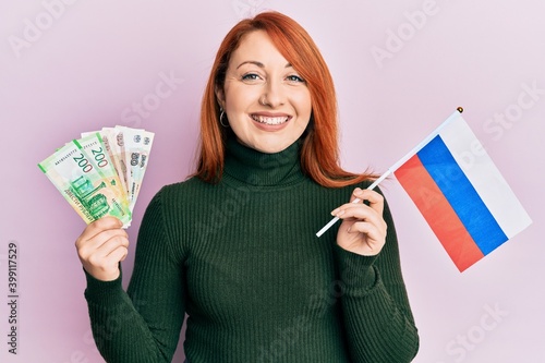 Beautiful redhead woman holding russian ruble banknotes and russia flag smiling with a happy and cool smile on face. showing teeth.