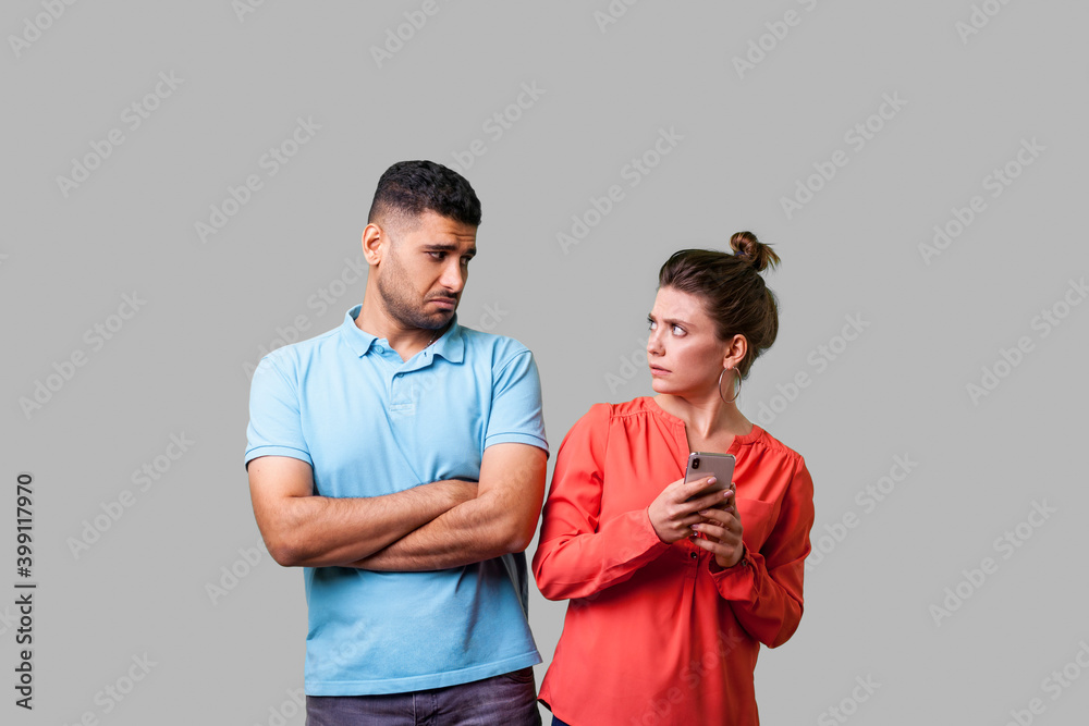 Portrait of funny resentful man in casual wear standing with crossed arms, looking disapprovingly at woman holding cellphone, addicted to internet. isolated on gray background, indoor studio shot Portrait of funny resentful man in casual wear standing with crossed arms, looking disapprovingly at woman holding cellphone, addicted to internet. isolated on gray background, indoor studio shot