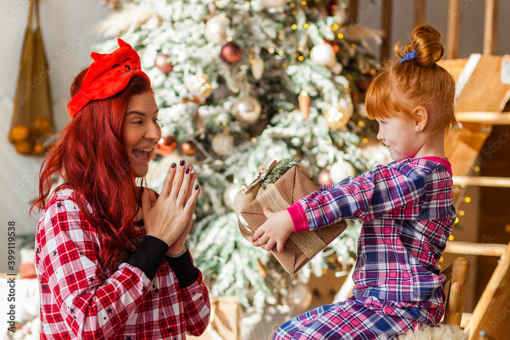 Happy woman and kid wearing checkered pajamas is sitting beside the Christmas tree open presents, winter holidays for mother and daughter