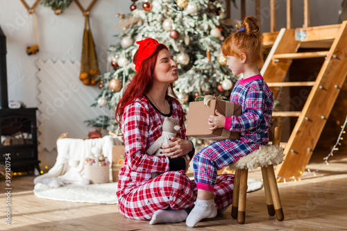 Happy woman and kid wearing checkered pajamas is sitting beside the Christmas tree open presents  winter holidays for mother and daughter