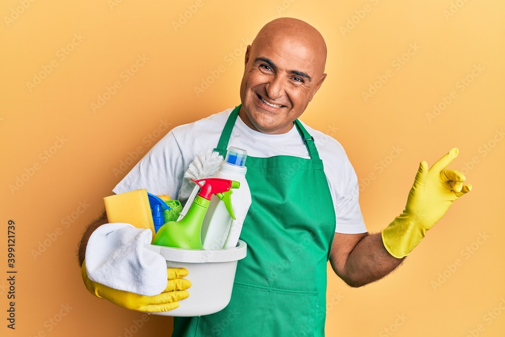 Mature middle east man wearing cleaner apron holding cleaning products smiling happy pointing with hand and finger to the side Mature middle east man wearing cleaner apron holding cleaning products smiling happy pointing with hand and finger to the side