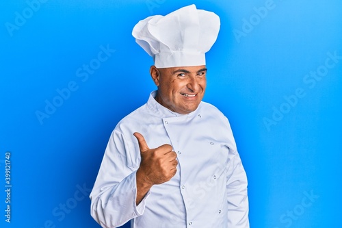 Mature middle east man wearing professional cook uniform and hat doing happy thumbs up gesture with hand. approving expression looking at the camera showing success.