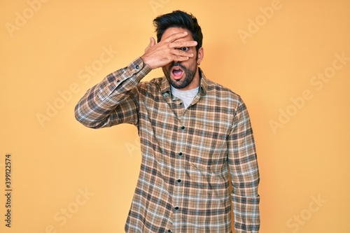 Handsome hispanic man with beard wearing casual clothes peeking in shock covering face and eyes with hand, looking through fingers with embarrassed expression.