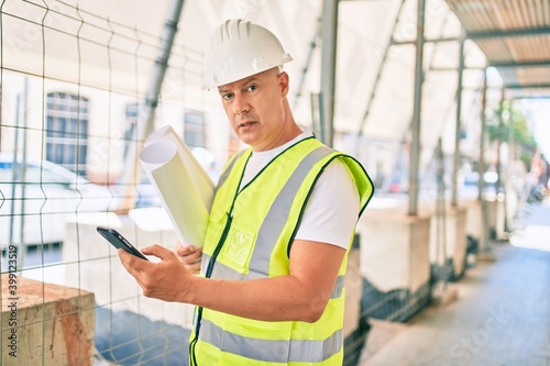 Middle age architect man with serious expression using smartphone and holding blueprints at the city.