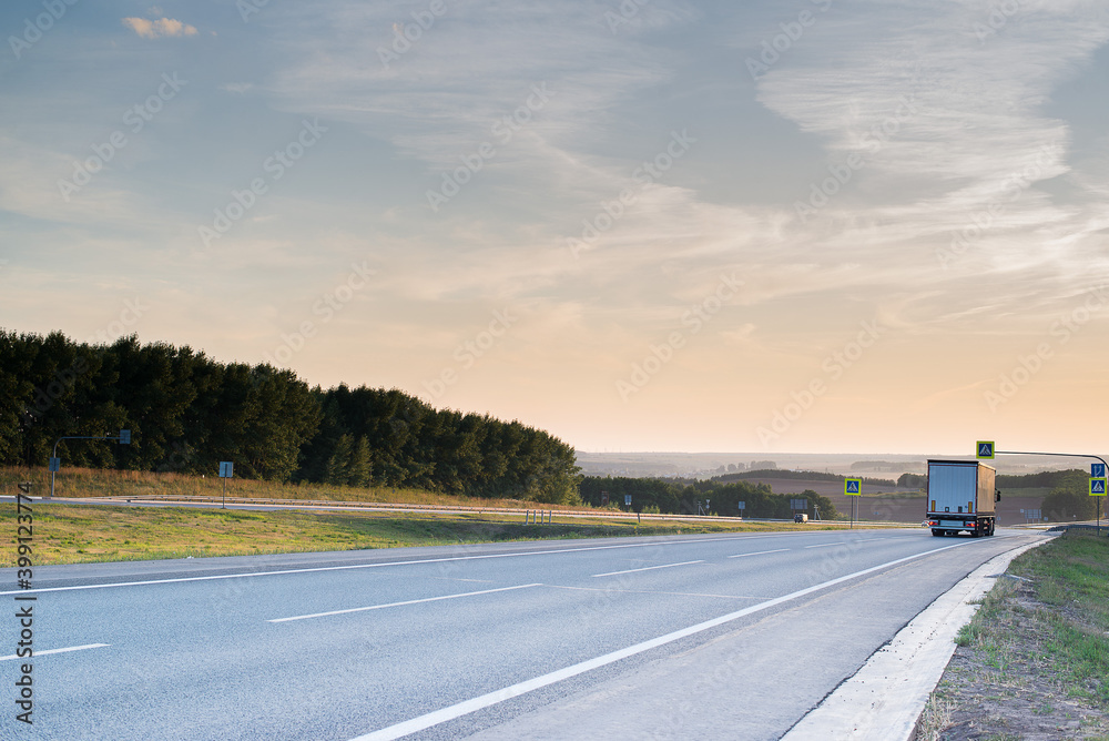 Fototapeta premium a cargo truck goes down a gray highway with a beautiful sunset in the sky and beautiful clouds in summer