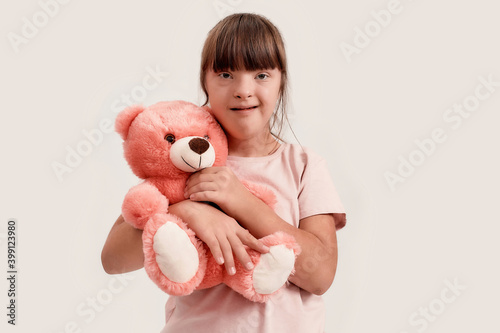 Portrait of disabled girl with Down syndrome looking at camera, holding her teddy bear while posing isolated over white background