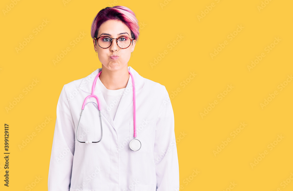 Young beautiful woman with pink hair wearing doctor uniform puffing cheeks with funny face. mouth inflated with air, crazy expression. Young beautiful woman with pink hair wearing doctor uniform puffing cheeks with funny face. mouth inflated with air, crazy expression.