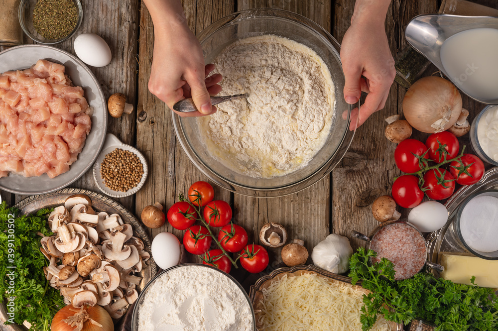Fototapeta premium Chef hands mix ingredients in glass bowl for cooking dough on wooden table with variety of ingredients background. Concept of cooking process. Backstage of preparing pie. View from above.