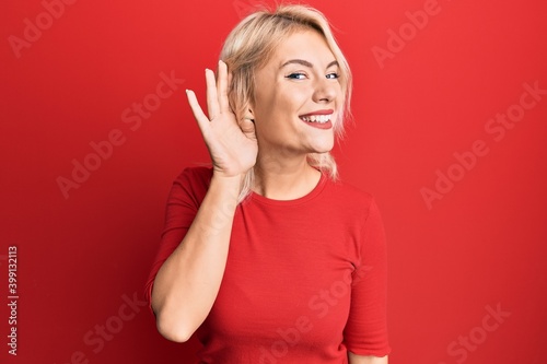 Young blonde girl wearing casual clothes smiling with hand over ear listening an hearing to rumor or gossip. deafness concept.