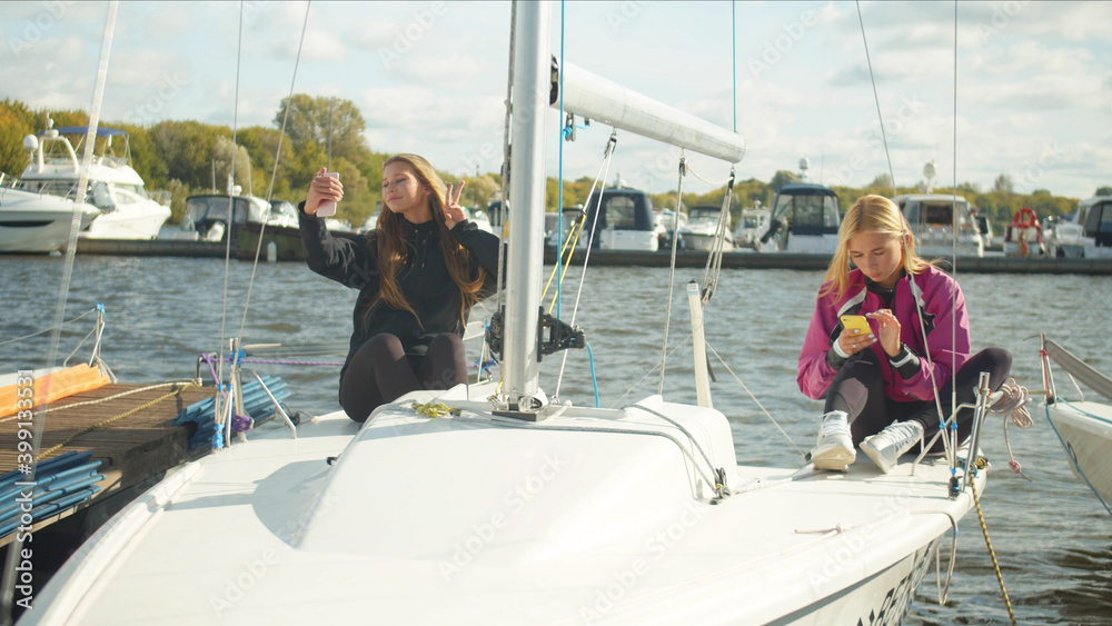 Young female sailing athletes sit on the deck of a sports yacht and take a selfie with a victory gesture.
