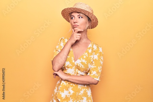 Beautiful blonde woman on vacation wearing summer hat and dress over yellow background with hand on chin thinking about question, pensive expression. Smiling with thoughtful face. Doubt concept.