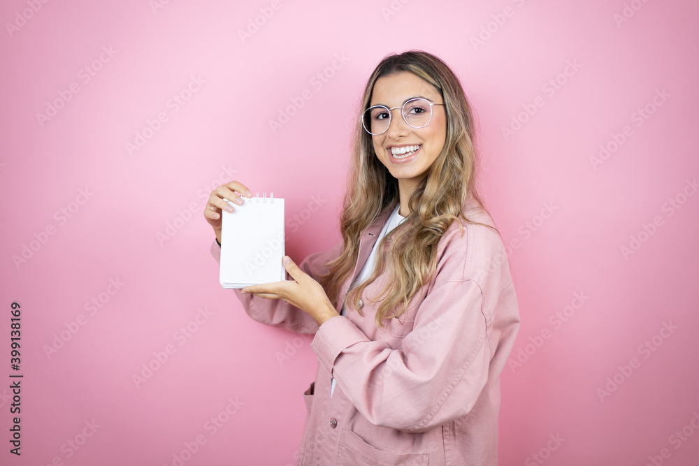Young beautiful blonde woman with long hair standing over pink background smiling and showing blank notebook in her hand Young beautiful blonde woman with long hair standing over pink background smiling and showing blank notebook in her hand