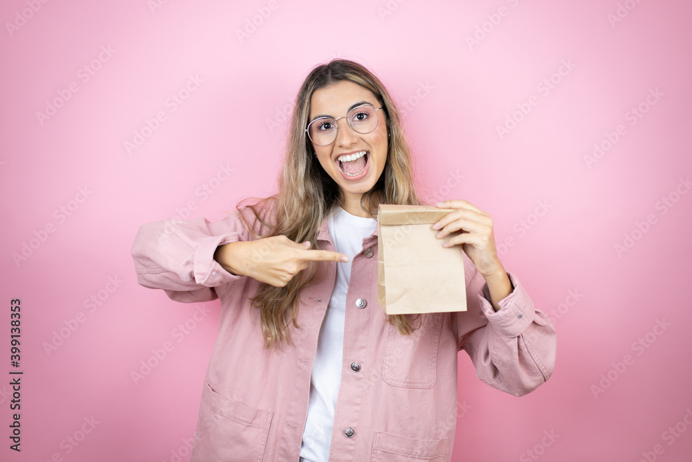 Young beautiful blonde woman with long hair standing over pink background smiling, surprised and pointing blank notebook in her hand Young beautiful blonde woman with long hair standing over pink background smiling, surprised and pointing blank notebook in her hand