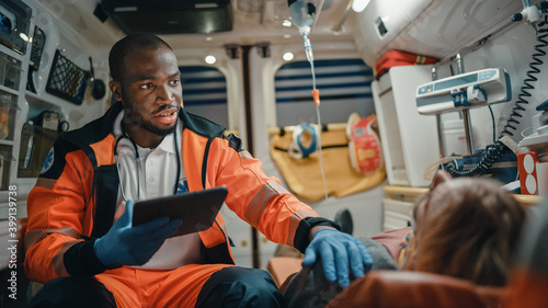 Black African American EMS Professional Paramedic Using Tablet Computer to Fill a Questionnaire for the Injured Patient on the Way to Hospital. Emergency Medical Care Assistant Works in an Ambulance.