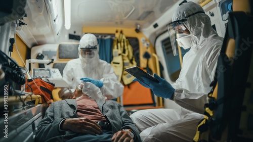 Paramedic in Coveralls Using Tablet Computer to Check the Diagnosis for the Injured Patient on the Way to Hospital. Emergency Medical Care Assistants Puts a Ventilation Mask in an Ambulance.