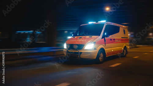 Parallel Shot of a Moving Ambulance Vehicle with Working Strobe Light and Signal Driving to Emergency Call on a City Urban Street at Night. Emergency Paramedics Rescue Van with Medical Cross Logo.