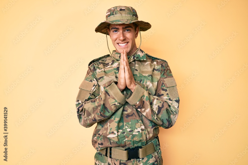 Young handsome man wearing camouflage army uniform praying with hands together asking for forgiveness smiling confident.