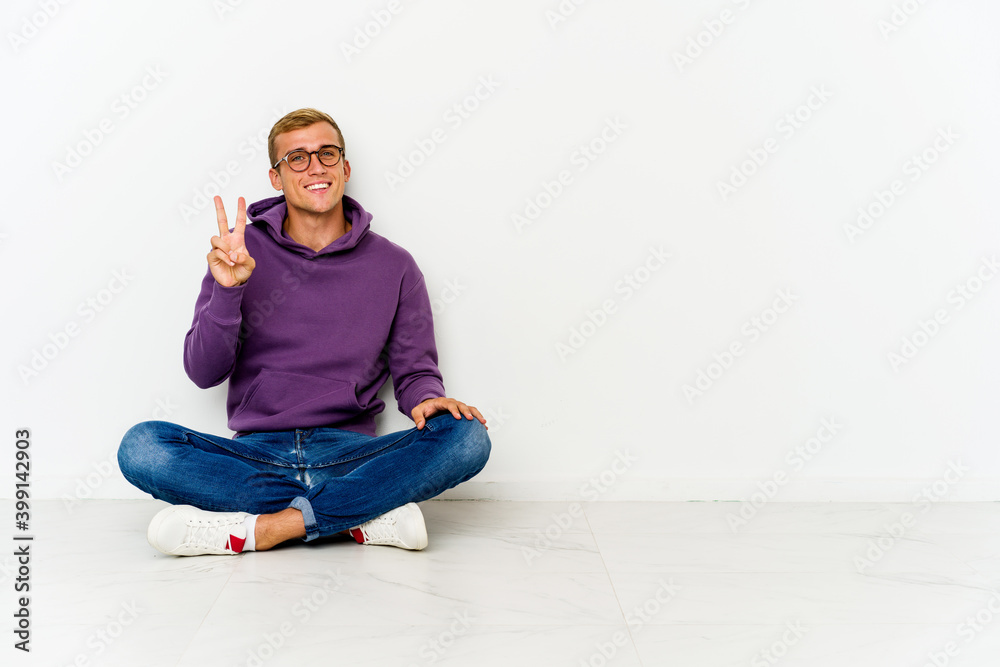 Young caucasian man sitting on the floor showing victory sign and smiling broadly. Young caucasian man sitting on the floor showing victory sign and smiling broadly.