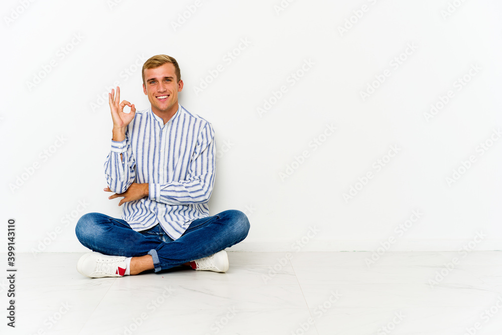 Young caucasian man sitting on the floor winks an eye and holds an okay gesture with hand. Young caucasian man sitting on the floor winks an eye and holds an okay gesture with hand.