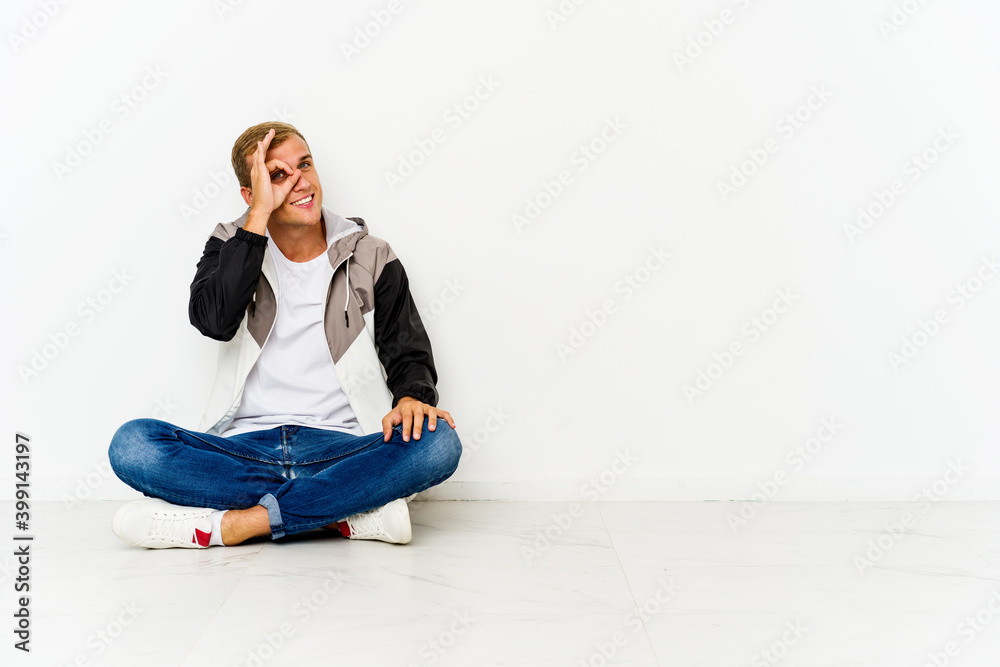 Young caucasian man sitting on the floor excited keeping ok gesture on eye. Young caucasian man sitting on the floor excited keeping ok gesture on eye.