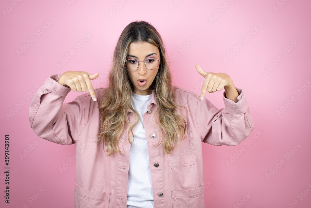 Young beautiful blonde woman with long hair standing over pink background surprised, looking down and pointing down with fingers and raised arms Young beautiful blonde woman with long hair standing over pink background surprised, looking down and pointing down with fingers and raised arms