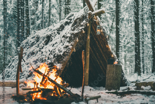 Primitive survival shelter in winter forest with Christmas garland near campfire