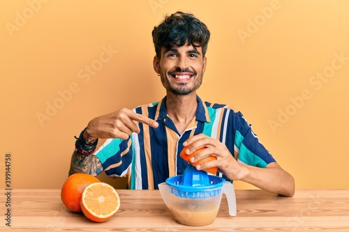 Young hispanic man sitting on the table using juicer smiling happy pointing with hand and finger