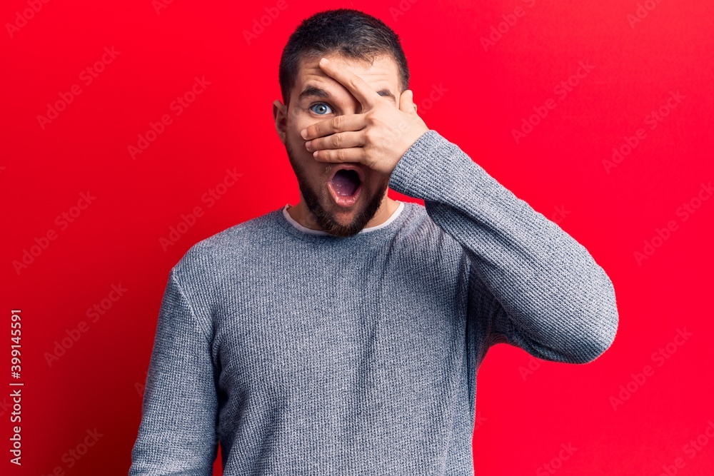 Young handsome man wearing casual sweater peeking in shock covering face and eyes with hand, looking through fingers afraid