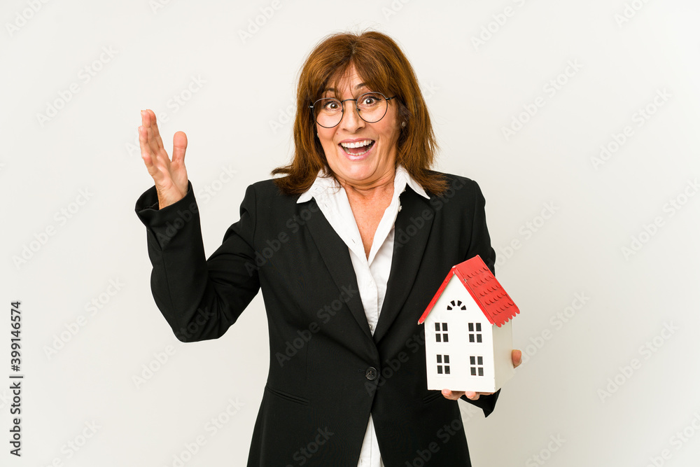 Middle aged real estate agent holding a house model isolated receiving a pleasant surprise, excited and raising hands. Middle aged real estate agent holding a house model isolated receiving a pleasant surprise, excited and raising hands.