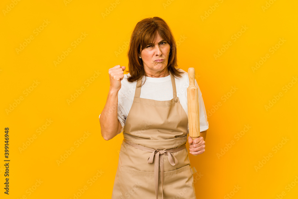 Middle age caucasian cook woman holding a roller isolated showing fist to camera, aggressive facial expression. Middle age caucasian cook woman holding a roller isolated showing fist to camera, aggressive facial expression.